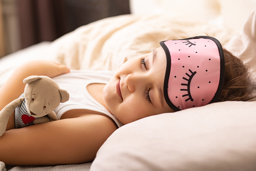 Cute little kid girl in a night mask lying on the bed and sleeping with teddy bear toy.
