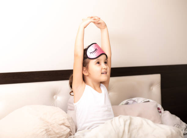 Cute little kid girl wake up in the morning, stretching hand rise up to the air while sitting in bed stock photo
