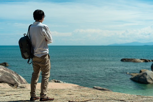 A young backpacker stands on the rocks by the sea, taking in a beautiful view. Traveling alone on vacation.