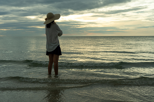 Woman wearing a hat walks alone on an empty sandy beach at sunset. A lonely and depressed young woman stands on the sand of the beach on holiday.