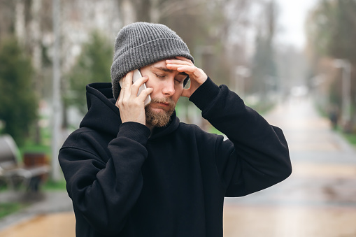 A bearded man in a hat is talking on the phone in the park and holding his head, bad news, problems and difficult questions.