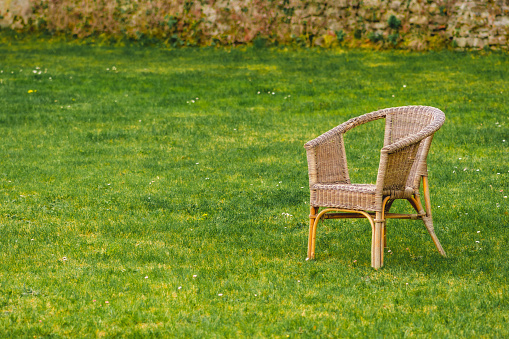 Old empty wicker chair on green grass lawn with flowers and old brick wall on background