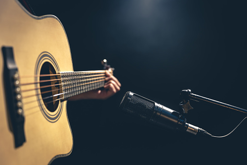 Male musician playing acoustic guitar behind microphone, close-up, recording in a music studio.