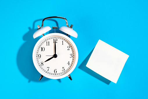 White alarm clock and blank white paper sticker on a blue background isolated, flat lay, copy space.