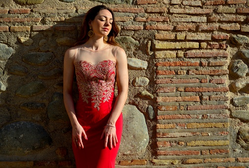 Portrait of young brunette woman in elegant oriental red dress against stone wall typical of Spain in summer, Europe