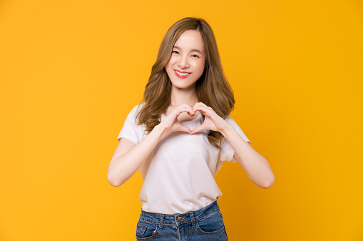 Studio shot of cheerful beautiful Asian woman in t-shirt and stand with showing heart on two hands on yellow background.