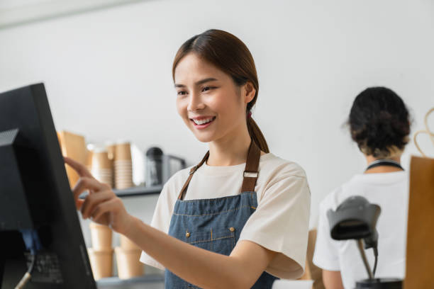 Happy young Asian woman cashier wears an apron and using pos terminal to input orders on coffee shop counter. stock photo
