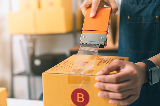 Startup small business, Woman owner using adhesive tape, packing carton parcel box working at the home office, seller prepares the delivery.