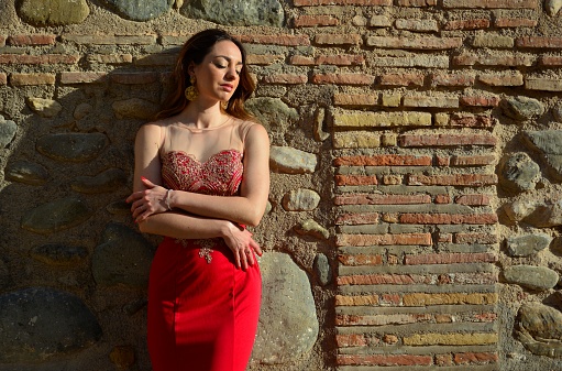 Portrait of young brunette woman in elegant oriental red dress against stone wall typical of Spain in summer, Europe