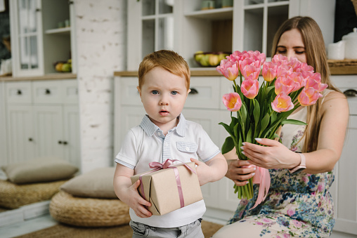 Mother's day concept. Cute little son congratulates mother and gives a bouquet of flowers tulips and a present box in the kitchen at home. Mom and child smiling and surprised. Family holiday together.