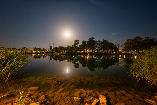 Night view of Balaton lake in Hungary, scenic landscape with yacht marine in moonlight, starry sky and reflection in the water, outdoor travel background