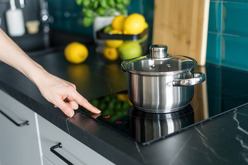 cropped shot of female hand press button on induction hob and choose temperature on control panel for cooking dinner in saucepan at home kitchen with stylish interior