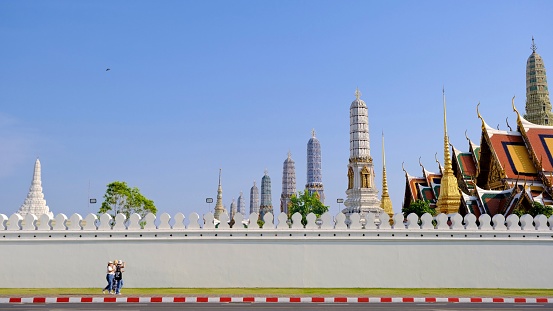 April-29-2023, Bangkok, Thailand :  \nTourists walk along the pathway of the Grand Palace with beautiful Stupas of Wat Phra Kaew behind the wall.