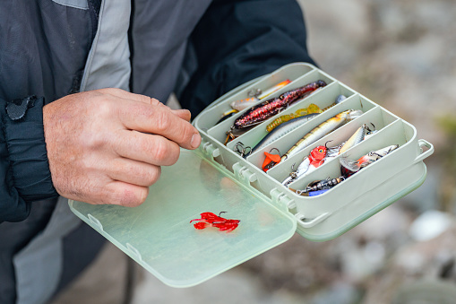 The fisherman's hands are holding a plastic box with a variety of lures and fishing hooks.