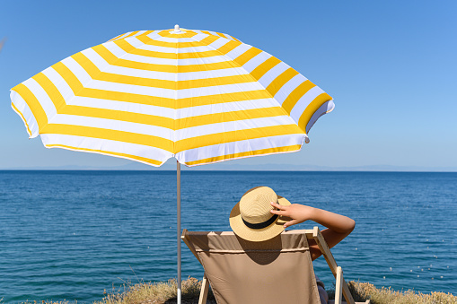 Woman in a straw hat sitting and sunbathing on sunbed, lounge, deck chair under yellow umbrella on the top of mountain, cliff with blue sea, ocean view.