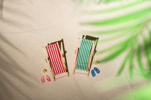 Aerial view two deck chair, glass of juice, flip flops on sand through the branches of palm and shadow palm tree. Travel, holiday, summertime, romantic concept. Top view.
