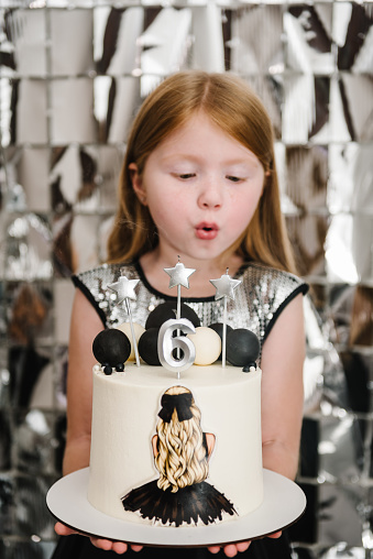 Girl holding birthday cake and blowing candles. Happy child celebrating birthday party 6 years. Photo wall, photo zone decoration white, black, silver balloons. Decor luxury style arch. Reception.