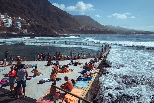 Bajamar, Tenerife, Spain, April 7, 2023: People bathing at the natural swimming pools on April 7, 2022 in Bajamar, Tenerife, Canary Islands,Spain. People chill out in the natural coastal pools while a rough sea hits the rocks of the harbor.