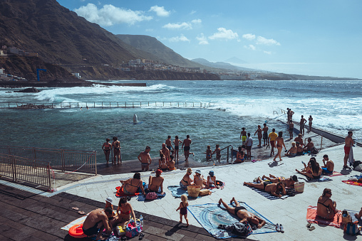 Bajamar, Tenerife, Spain, April 7, 2023: People bathing at the natural swimming pools on April 7, 2022 in Bajamar, Tenerife, Canary Islands,Spain. People chill out in the natural coastal pools while a rough sea hits the rocks of the harbor.