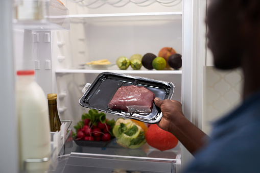 Close up black man taking piece of meat out of refrigerator while cooking dinner at home