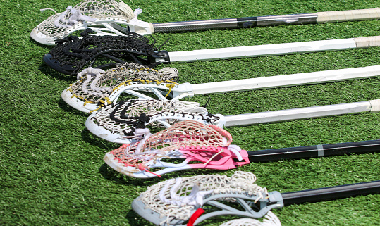 Six colorful lacrosse sticks lying on a green turf field in a row.