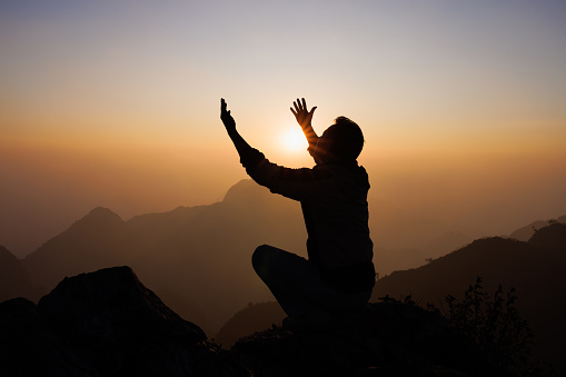 Silhouette of a man is praying to God on the mountain. Praying hands with faith in religion and belief in God on blessing background. Power of hope or love and devotion.