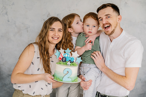 Happy family holds cake and having fun celebrating a birthday party 2 years. Mom, dad, daughter congratulate son on birthday near grey wall in studio. Mother, father hugging kids. Birthday reception.