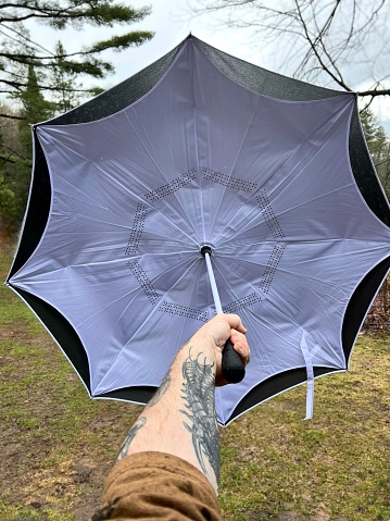 Tattooed man’s arm holding an umbrella up in the spring rain. Taken on a hobby farm in Northern Wisconsin. Barely turning green yard, forest, sky, and tree branches can be seen around the large umbrella.
