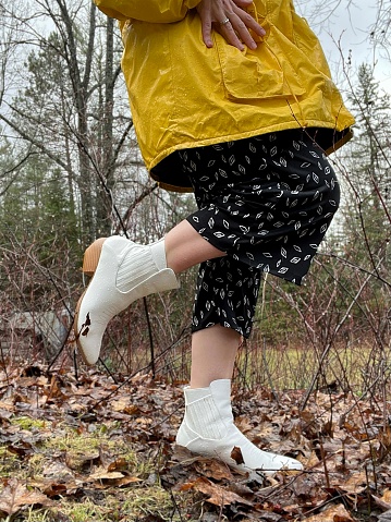 Partial lower view of a woman outdoors enjoying the spring rain in her yellow rain jacket. Photo is taken from the waist down only, part of her raincoat, pants, and white cowboy boots are shown in the photo. Taken in a rural yard in Northern Wisconsin with grass and trees surrounding the woman.