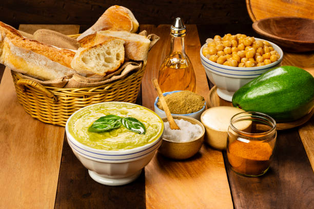 View of a white bowl with hummus made of chick peas and avocado and basil leaves and chilly red pepper powder over it. Also with ingredients on the table are: Ingredients in the photo: bread to be spread with the hummus, extra virgin olive oil, red chilly pepper powder, salt, sesame cream and cooked chick peas ,oregano and avocado.