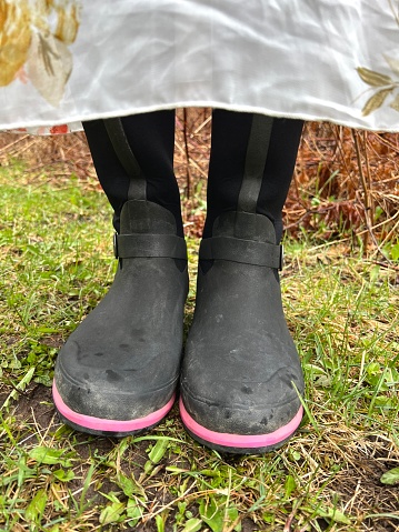 Partial lower view of a teenagers rubber boots and flowered dress. Only a small part of her dress can be seen, she is lifting it above her boots, standing in her emerging spring yard. Taken in a rural yard in Northern Wisconsin with grass and plants surrounding the teen.