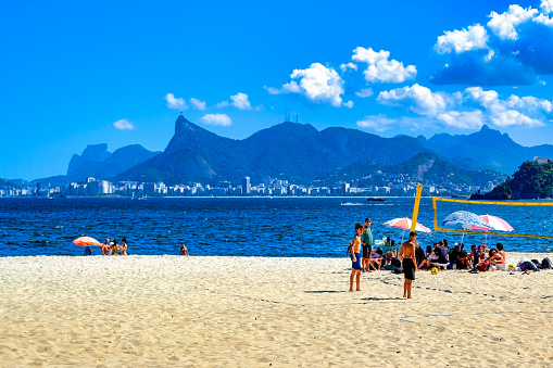 Niteroi, Rio de Janeiro, Brazil - April 21, 2023: Two boys stand beside a volleyball net on a sunny sandy beach with a group of people relaxing under canopies. The ocean, cityscape, mountains, and blue sky are visible in the background.