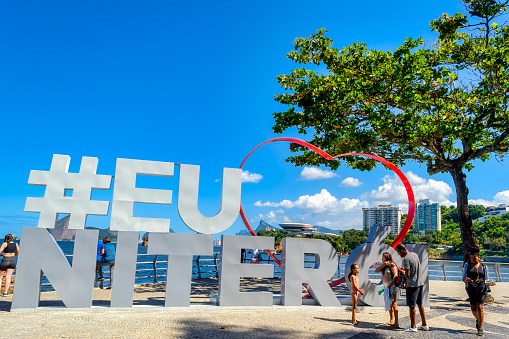 Niteroi, Rio de Janeiro, Brazil - April 21, 2023: The words “#EU NITEROI” in large letters on a sandy beach, with a large red heart towering above and partially resting on it. People are standing close to the sign in the foreground, and the sea, blue sky, and buildings are visible in the background.