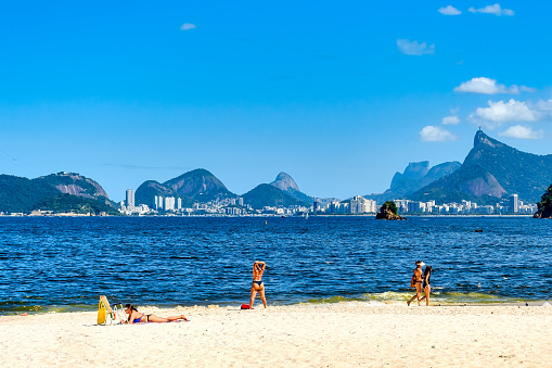 Niteroi, Rio de Janeiro, Brazil - April 21, 2023: People walking and relaxing on a sandy beach on a sunny day. A cityscape, mountainscape, and the blue sky are visible across the sea.