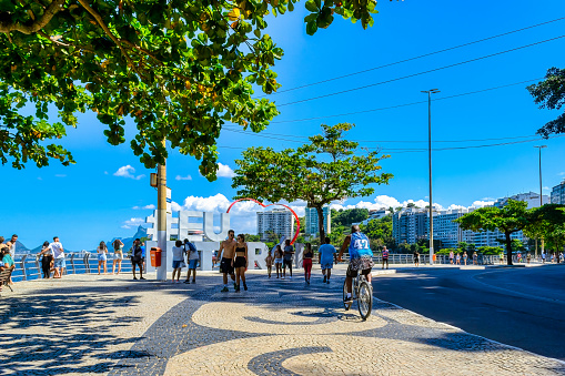 Niteroi, Rio de Janeiro, Brazil - April 21, 2023: A city street with people walking, cycling, and standing on the sidewalk. Trees line the sides of the street, and skyscrapers and the blue sea are visible in the distance.