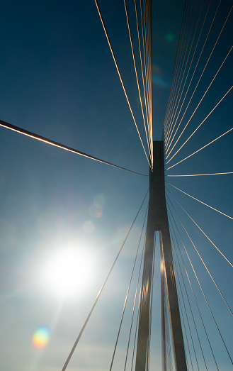 Support and cables of the suspension bridge against the background of the blue sky and the glare of the sunbeam. Buildings and structures in Norway. Vertical photo, bottom view