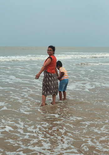 Mandarmani, 04-22-2023: a young boy holding hands of her mother looking at horizon over sea. standing in knee deep water on beach. The woman is looking back and smiling at photographer. Mandarmani is a popular fishing village and seaside travel destination.