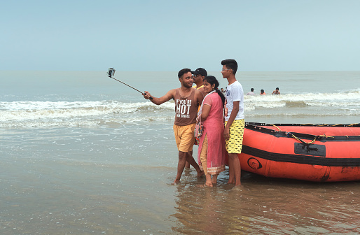Mandarmani, 04-22-2023: happy tourists taking selfie in front of an inflatable rubber boat, during their visit on the beach. Splashing sea waves along the shore are seen in background.