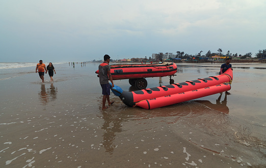 Mandarmani, 04-22-2023: A young couple is seen walking across tour boat operators carrying an inflatable rubber boats (taking tourists for thrilling water ride) towards sea, against backdrop of an overcast sky in a monsoon day.