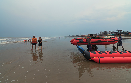 Mandarmani, 04-22-2023: A young couple is seen walking across tour boat operators carrying an inflatable rubber boats (taking tourists for thrilling water ride) towards sea, against backdrop of an overcast sky in a monsoon day.