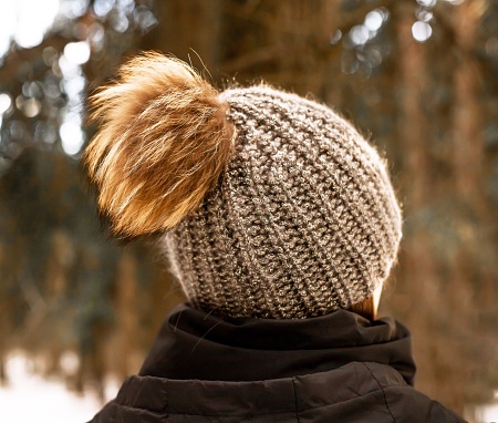 Knitted hat with fluffy pompom, back side view, on female head outdoors in winter nature.