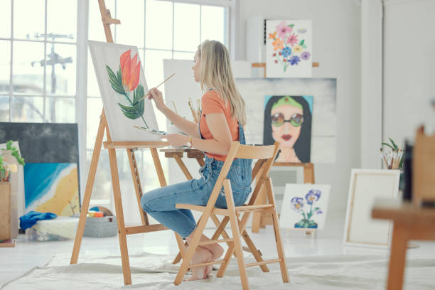 istock Paint, art or flower with a woman in a studio or workshop painting on a canvas using an easel for expression. Watercolor, artistic and nature with a female painter sitting in her home from the back 1486863815