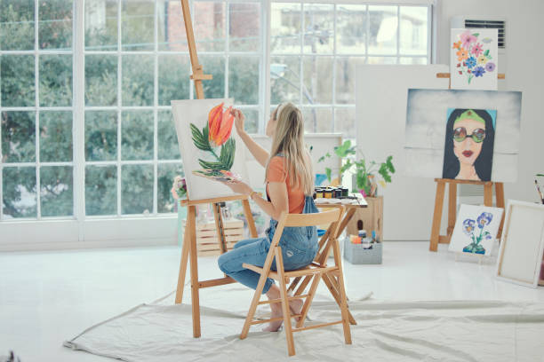 istock Painting, art or flower with a woman in a studio or workshop adding color to canvas using an easel and oil paint. Watercolor, artistic and creative with a young female painter sitting in her home 1486863817