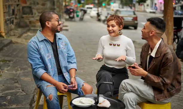 Three diverse young friends smiling and talking together while having a bite to eat at a sidewalk cafe