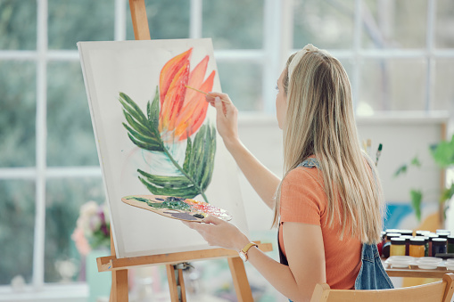 Painter, flower art or creative with a woman in a studio or workshop painting on a canvas using an easel and oil paint. Watercolor, inspiration and back with a young female artist sitting in her home