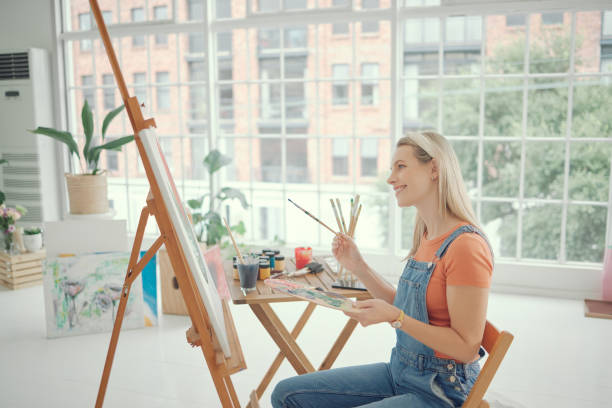 istock Paint, art and happy with a woman in a studio or workshop painting on a canvas using an easel and watercolor. Oil color, inspiration and artistic with a young female painter sitting in her home 1486866145