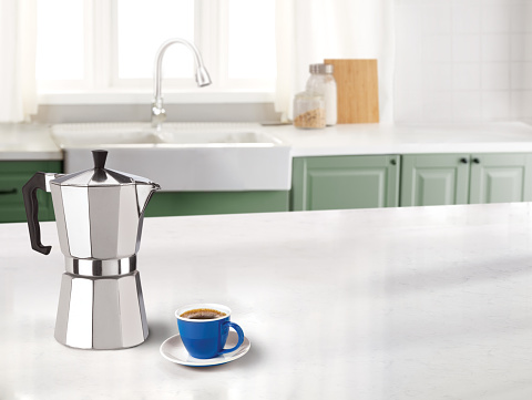 Stovetop espresso maker with a coffee cup in a kitchen on a marble counter top and sink in the background
