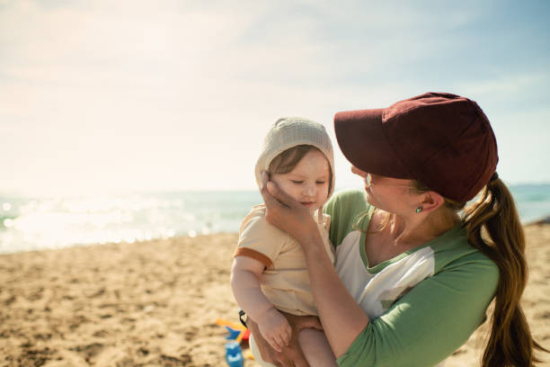 Woman applying sun protection lotion to her baby girl near the sea on beach holidays stock photo