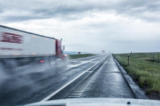 Somewhere on route Interstate 80 in the western USA state of Wyoming, a speeding semi tractor trailer truck splashes and kicks up foggy water and sloppy haze as it zooms past a stationary car parked on the highway shoulder during an extreme weather rain storm on the wilderness prairie.