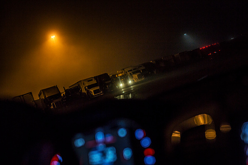 View through a parked car windshield toward a very tall yellow-orange high pressure sodium vapor street light which is cutting through a stormy overcast foggy night above an interstate expressway trailer truck rest stop with dozens of freight transportation vehicles parked in rows. One truck has its headlights on - ready to pull out and get back onto the midnight highway.